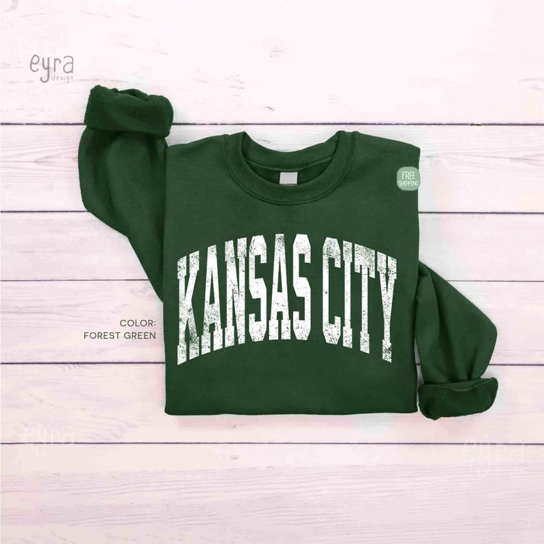 May include: A forest green sweatshirt featuring the text "KANSAS CITY" in distressed white lettering. The sweatshirt has a crew neck and cuffed sleeves. The colour is forest green.