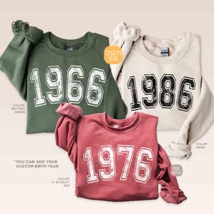 May include: Three sweatshirts in military green, sand, and H. scarlet red, each displaying a year in a distressed white font. The military green sweatshirt shows "1966", the sand one "1986", and the red one "1976". The text "CUSTOM YEAR" is also visible.