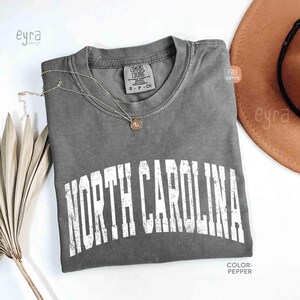 K&ouml;nnte beinhalten: Ein pfefferfarbenes Kurzarm-T-Shirt mit dem Schriftzug NORTH CAROLINA in verwaschener wei&szlig;er Schrift auf der Brust. Das Shirt hat einen Rundhalsausschnitt und ein Comfort Colors Markenetikett am Innenkragen. Eine d&uuml;nne goldfarbene Kette liegt darauf.