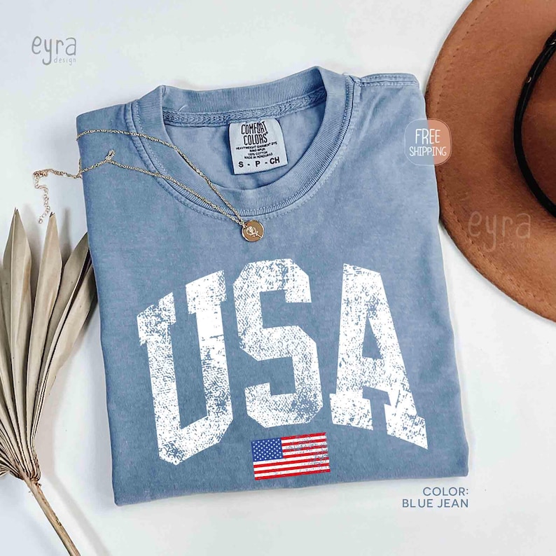 Puede incluir: Una camiseta azul vaquero con la palabra "USA" en letras blancas grandes y desgastadas. Debajo de las letras hay una peque&ntilde;a bandera americana. Un collar dorado descansa sobre la camisa. La camiseta es de la marca Comfort Colors.
