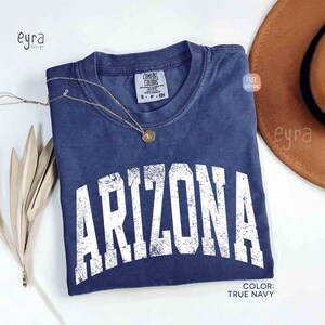 May include: A true navy blue short-sleeve t-shirt with a crew neckline and a distressed white graphic print that reads ARIZONA in a large, arched, collegiate-style font. The shirt features a Comfort Colors brand label at the inner neck.