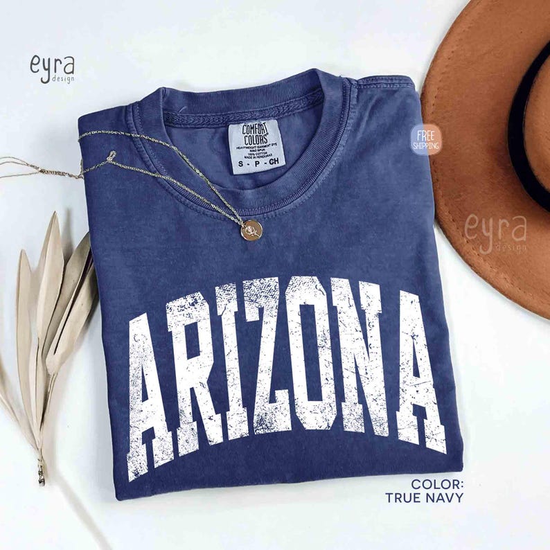 May include: A true navy blue short-sleeve t-shirt with a crew neckline and a distressed white graphic print that reads ARIZONA in a large, arched, collegiate-style font. The shirt features a Comfort Colors brand label at the inner neck.
