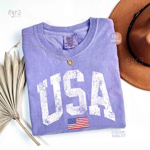 Puede incluir: Una camiseta de color violeta con la palabra "USA" en letras blancas grandes y desgastadas. Debajo del texto hay una peque&ntilde;a bandera americana. Tambi&eacute;n hay un collar dorado y un sombrero marr&oacute;n en la imagen.