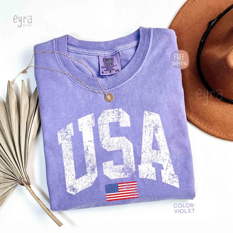 Puede incluir: Una camiseta de color violeta con la palabra "USA" en letras blancas grandes y desgastadas. Debajo del texto hay una peque&ntilde;a bandera americana. Tambi&eacute;n hay un collar dorado y un sombrero marr&oacute;n en la imagen.