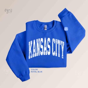 May include: Royal blue sweatshirt with the text "KANSAS CITY" in distressed white lettering. The sweatshirt has a crew neck and long sleeves. The colour is described as Royal Blue.