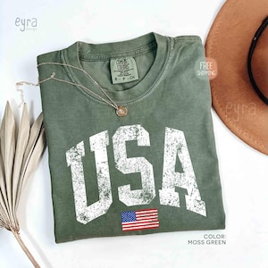 Puede incluir: Camiseta verde musgo con la palabra "USA" en letras blancas desgastadas y una peque&ntilde;a bandera americana. Un collar dorado y un sombrero marr&oacute;n est&aacute;n junto a la camiseta. La camiseta est&aacute; etiquetada como "Comfort Colors" y es talla S-P-CH.