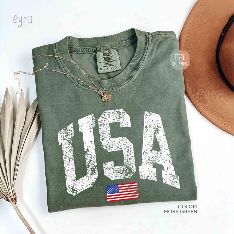 Puede incluir: Camiseta verde musgo con la palabra "USA" en letras blancas desgastadas y una peque&ntilde;a bandera americana. Un collar dorado y un sombrero marr&oacute;n est&aacute;n junto a la camiseta. La camiseta est&aacute; etiquetada como "Comfort Colors" y es talla S-P-CH.