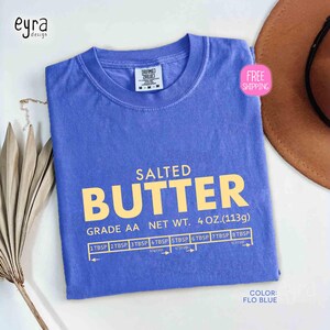 May include: A Flo Blue t-shirt with the words "SALTED BUTTER" in large yellow letters. Below the text are measurements in tablespoons and cups. The shirt also has the text "GRADE AA NET WT. 4 OZ. (113g)".