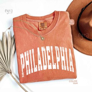 May include: A terracotta-coloured short-sleeve t-shirt featuring the word PHILADELPHIA printed in a distressed white collegiate font across the chest. The shirt has a crew neckline and a Comfort Colors brand label inside the collar.