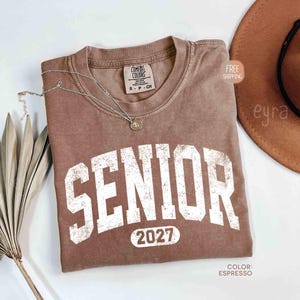 May include: Espresso-colored t-shirt with the word "SENIOR" in large, distressed white letters and the year "2027" below. The shirt has a crew neck and a small tag. A silver necklace with a pendant is draped across the shirt. A brown hat is in the background.