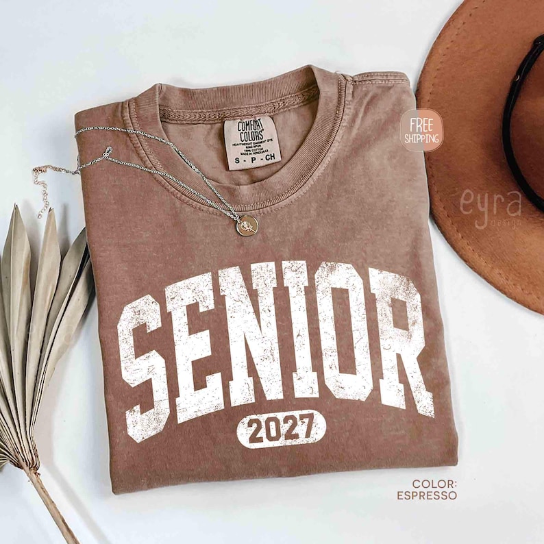 May include: Espresso-colored t-shirt with the word "SENIOR" in large, distressed white letters and the year "2027" below. The shirt has a crew neck and a small tag. A silver necklace with a pendant is draped across the shirt. A brown hat is in the background.