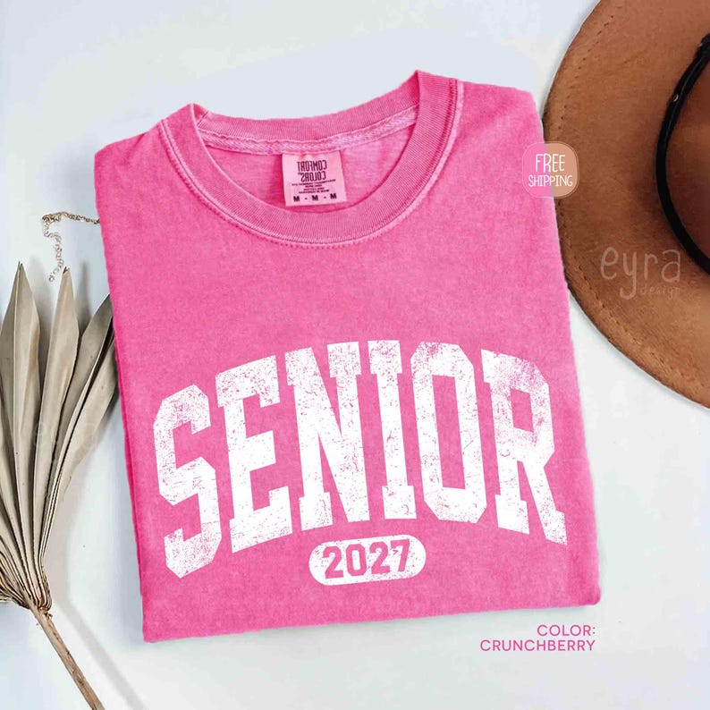 May include: A pink t-shirt with the word "SENIOR" in large white, distressed letters. Below the word "SENIOR" is the number "2027". The shirt is a solid color and has a crew neck. The color is "Crunchberry".