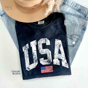 Puede incluir: Camiseta negra con la palabra "USA" en letras blancas desgastadas y un peque&ntilde;o parche de la bandera estadounidense. La camiseta se combina con vaqueros de mezclilla de lavado claro y un sombrero beige.