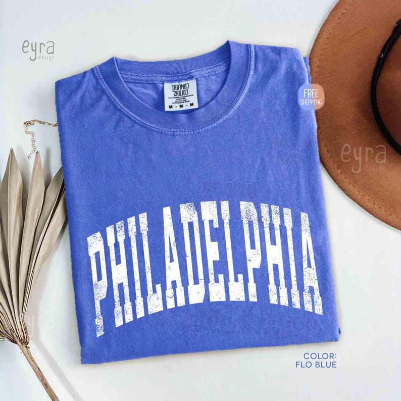 May include: A periwinkle blue short-sleeve t-shirt with a crew neckline. The shirt features the word PHILADELPHIA printed in a large, arched, distressed white font across the chest. A white tag inside the collar reads Comfort Colors size M.