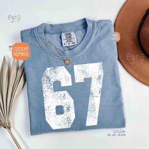 K&ouml;nnte beinhalten: Ein blaues Jeans-T-Shirt mit der Nummer 67 in wei&szlig;er, verblichener Schrift. Das Shirt hat einen Rundhalsausschnitt und ein Etikett mit der Aufschrift "Comfort Colors". Eine goldene Halskette mit Anh&auml;nger befindet sich auf dem Shirt. Das Shirt ist Gr&ouml;&szlig;e small.