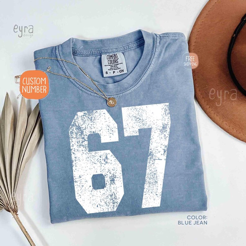 K&ouml;nnte beinhalten: Ein blaues Jeans-T-Shirt mit der Nummer 67 in wei&szlig;er, verblichener Schrift. Das Shirt hat einen Rundhalsausschnitt und ein Etikett mit der Aufschrift "Comfort Colors". Eine goldene Halskette mit Anh&auml;nger befindet sich auf dem Shirt. Das Shirt ist Gr&ouml;&szlig;e small.