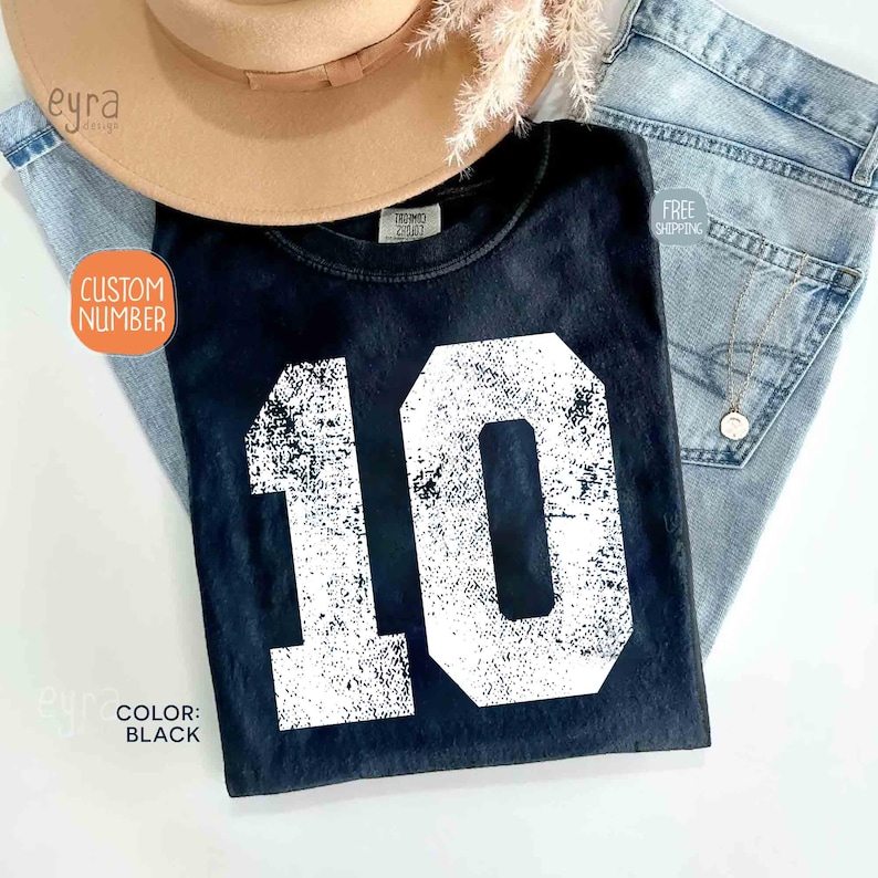 K&ouml;nnte beinhalten: Schwarzes T-Shirt mit der wei&szlig;en Zahl 10 in verblichener Schrift. Das Shirt wird mit einem beigefarbenen Hut und einer blauen Jeans pr&auml;sentiert. Ein orangefarbener Kreis zeigt "CUSTOM NUMBER". Der Text "COLOR: BLACK" ist ebenfalls sichtbar.