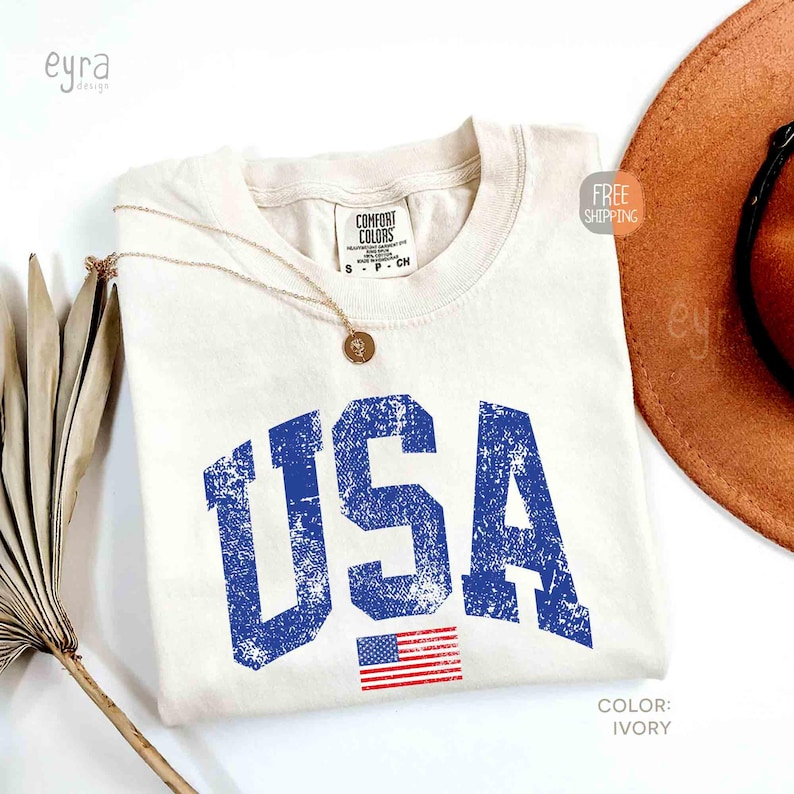 Puede incluir: Camiseta de color marfil con un gr&aacute;fico azul desgastado de "USA" y una peque&ntilde;a bandera estadounidense. Un collar dorado con un colgante descansa sobre la camiseta. Un sombrero marr&oacute;n es parcialmente visible en el fondo.