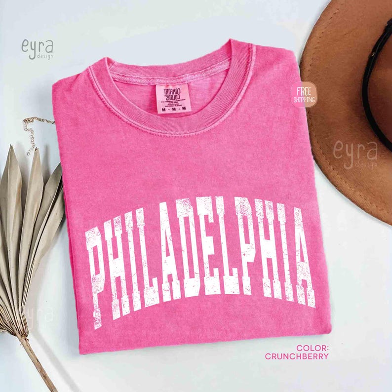 May include: A short-sleeve crew neck t-shirt in a bright pink colour with a distressed white arched graphic that reads PHILADELPHIA across the chest. The shirt features a Comfort Colors brand label at the inner neckline and a size M tag.