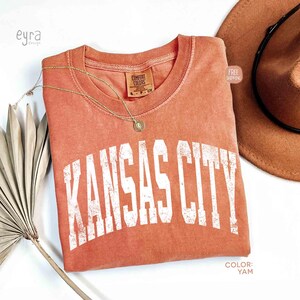 May include: A coral-colored t-shirt with the text "KANSAS CITY" in white, distressed lettering. A gold necklace and a brown hat are also visible. The shirt has a Comfort Colors tag.