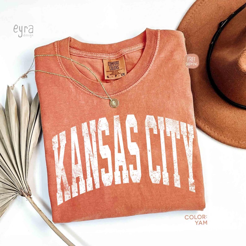 May include: A coral-colored t-shirt with the text "KANSAS CITY" in white, distressed lettering. A gold necklace and a brown hat are also visible. The shirt has a Comfort Colors tag.