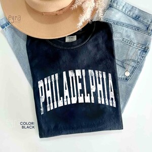 May include: A black t-shirt with a distressed white arched Philadelphia graphic across the chest. The shirt is layered with a tan wide-brim hat and light blue denim jeans. A small gold necklace with a circular pendant rests on the denim fabric.
