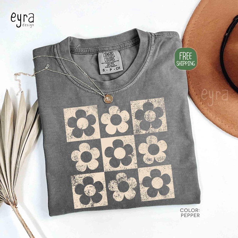 May include: A gray Comfort Colors t-shirt with a grid of nine beige flowers. The shirt has a crew neck and short sleeves. A silver necklace with a pendant rests on the shirt. The color of the shirt is pepper.