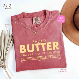 May include: Crimson-colored t-shirt with the text "SALTED BUTTER" in yellow. The shirt includes a graphic with measurements and the text "GRADE AA NET WT. 4 OZ. (113g)". A silver necklace with a pendant is on the shirt.