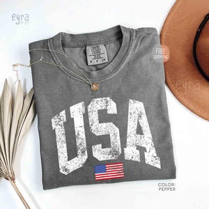 Puede incluir: Una camiseta gris Comfort Colors con la palabra "USA" en letras blancas desgastadas y una peque&ntilde;a bandera estadounidense. Tambi&eacute;n se ven un collar plateado y un sombrero marr&oacute;n.