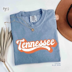 May include: A blue jean coloured t-shirt with the word "Tennessee" in white text with an orange outline. The shirt has a crew neck and a small tag at the neckline. A gold necklace with a pendant rests on the shirt.