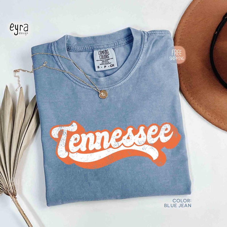 May include: A blue jean coloured t-shirt with the word "Tennessee" in white text with an orange outline. The shirt has a crew neck and a small tag at the neckline. A gold necklace with a pendant rests on the shirt.