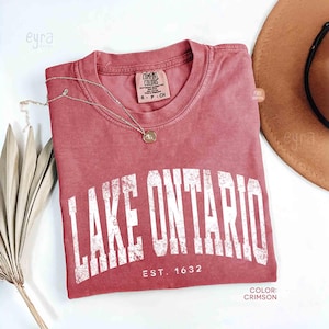 Puede incluir: Una camiseta de manga corta color carmesí con cuello redondo que presenta el texto blanco desgastado "LAKE ONTARIO EST. 1632" impreso en el pecho. La prenda incluye una etiqueta de la marca Comfort Colors en el cuello y un collar dorado fino.