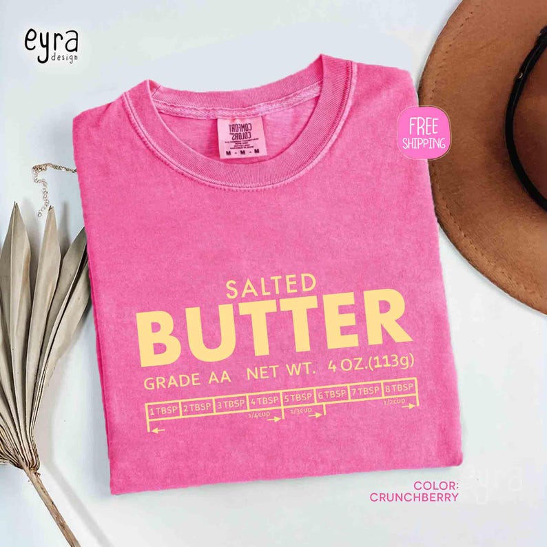 May include: A pink t-shirt with the text "SALTED BUTTER" in yellow. The shirt also features the text "GRADE AA NET WT. 4 OZ. (113g)" and a measurement guide. The shirt's color is "CRUNCHBERRY".