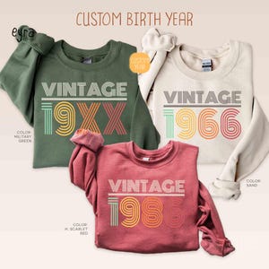 Pode incluir: Três camisolas de gola redonda em verde militar, areia e vermelho H. escarlate. Cada uma apresenta a palavra "VINTAGE" acima de um ano em uma fonte retrô. O texto "CUSTOM BIRTH YEAR" está no topo. A camisola verde militar diz "19XX", a camisola areia diz "1966" e a camisola vermelha diz "1986".