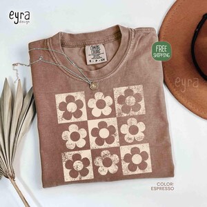 May include: Espresso-colored t-shirt with a grid of cream-colored flowers. The shirt has a crew neck and short sleeves. A silver necklace with a pendant is draped on the shirt. The tag reads "COMFORT COLORS".