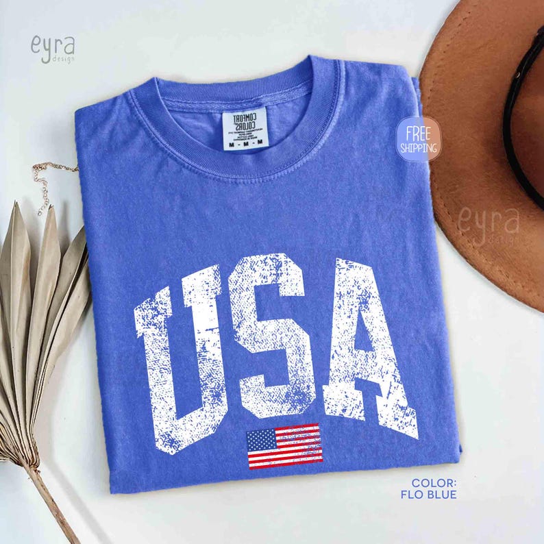 Puede incluir: Una camiseta azul con la palabra "USA" en letras blancas grandes y desgastadas. Debajo del texto hay una peque&ntilde;a bandera estadounidense. La camiseta es de talla mediana y de color azul Flo.