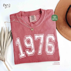 May include: Crimson-colored t-shirt with the number "1976" printed in white. The shirt has a crew neck and a small tag on the collar. A gold necklace with a pendant is draped on the shirt. The shirt is labeled "Comfort Colors".