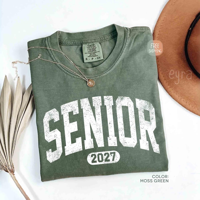 May include: Moss green t-shirt with the word "SENIOR" in distressed white lettering and the year "2027" below. A gold-colored necklace with a pendant rests on the shirt. The shirt is a Comfort Colors brand.