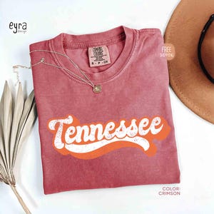 May include: Crimson t-shirt featuring the word "Tennessee" in white and orange retro lettering. The shirt has a crew neck and short sleeves. A gold necklace with a pendant rests on the shirt. A brown hat is in the background.