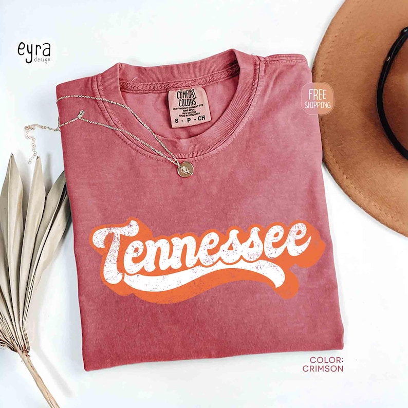 May include: Crimson t-shirt featuring the word "Tennessee" in white and orange retro lettering. The shirt has a crew neck and short sleeves. A gold necklace with a pendant rests on the shirt. A brown hat is in the background.