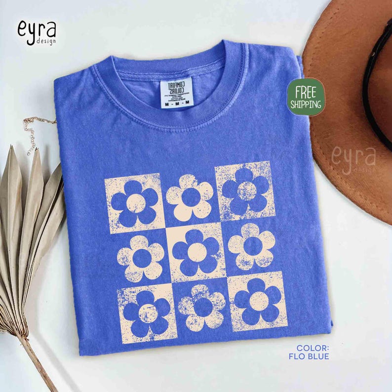 May include: A blue t-shirt with a retro floral design. The shirt features a grid of nine flowers, each in a square. The flowers are light beige with blue centers. The shirt is a solid blue color.