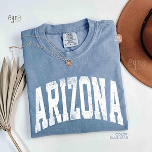 May include: A blue jean colored short-sleeve t-shirt with a crew neckline. The shirt features the word ARIZONA printed in a large, distressed white collegiate-style font across the chest. A gold-toned necklace with a small circular pendant rests on the collar.