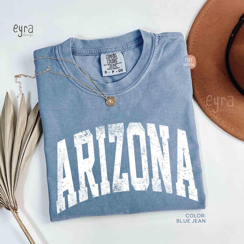 May include: A blue jean colored short-sleeve t-shirt with a crew neckline. The shirt features the word ARIZONA printed in a large, distressed white collegiate-style font across the chest. A gold-toned necklace with a small circular pendant rests on the collar.