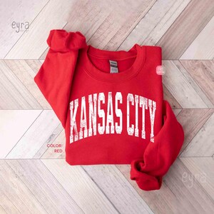 May include: A red sweatshirt with the text "KANSAS CITY" in white, distressed lettering. The sweatshirt is folded on a light wood-grain surface. The colour is indicated as red.