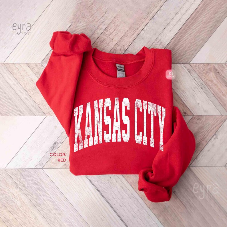 May include: A red sweatshirt with the text "KANSAS CITY" in white, distressed lettering. The sweatshirt is folded on a light wood-grain surface. The colour is indicated as red.