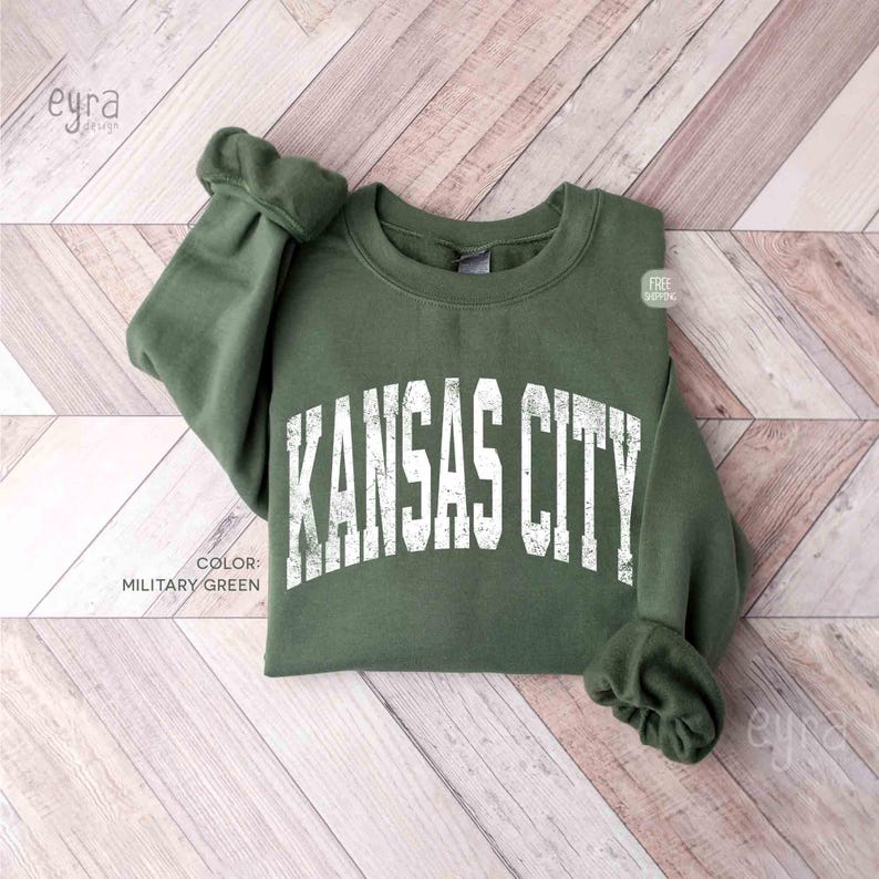 May include: A military green sweatshirt featuring the text "KANSAS CITY" in white, distressed lettering. The garment is displayed on a light wood-effect surface. The colour is described as "MILITARY GREEN".