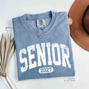 May include: A blue jean-colored t-shirt with the word "SENIOR" in large, distressed white letters. Below the word is the number "2027". A gold necklace with a pendant rests on the shirt. The shirt is a Comfort Colors brand.
