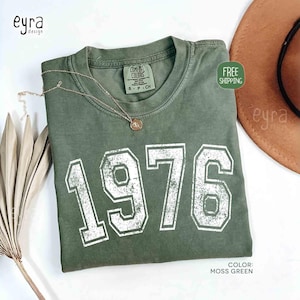 May include: A moss green t-shirt with the year "1976" printed in white, distressed lettering. A gold-colored necklace with a pendant rests on the shirt. The shirt is folded and displayed with a brown hat.