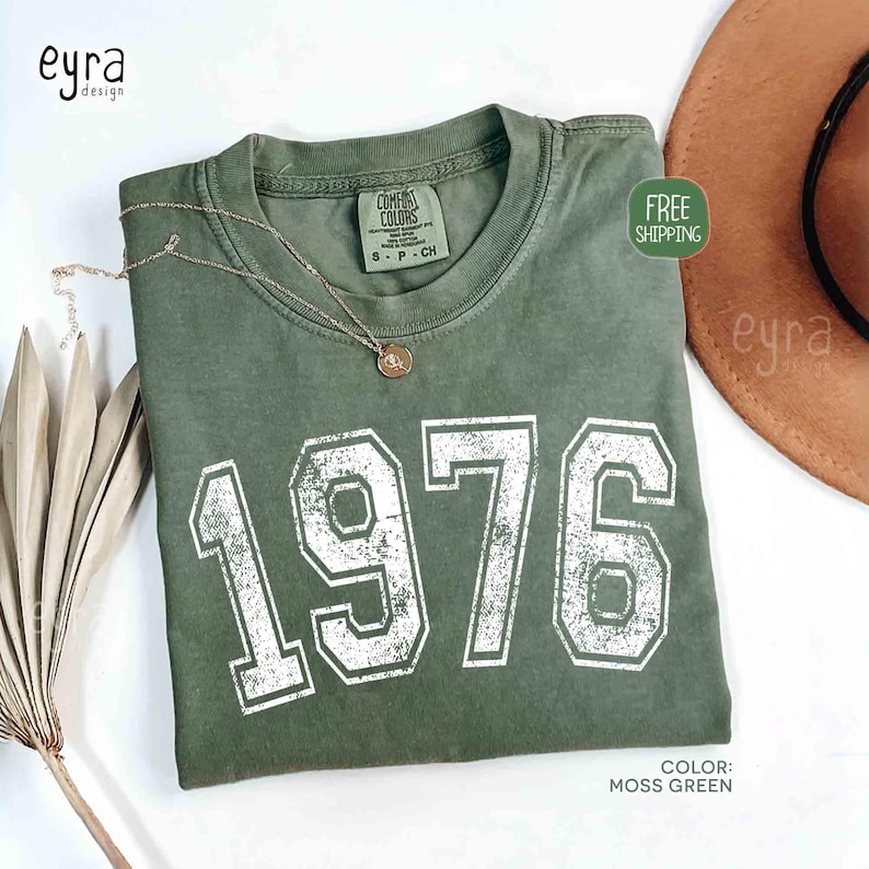 May include: A moss green t-shirt with the year "1976" printed in white, distressed lettering. A gold-colored necklace with a pendant rests on the shirt. The shirt is folded and displayed with a brown hat.