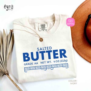 May include: Ivory-colored t-shirt with the words "SALTED BUTTER" in blue, along with measurement conversions. The shirt also features the text "GRADE AA NET WT. 4 OZ. (113g)". A gold necklace with a pendant is draped on the shirt.